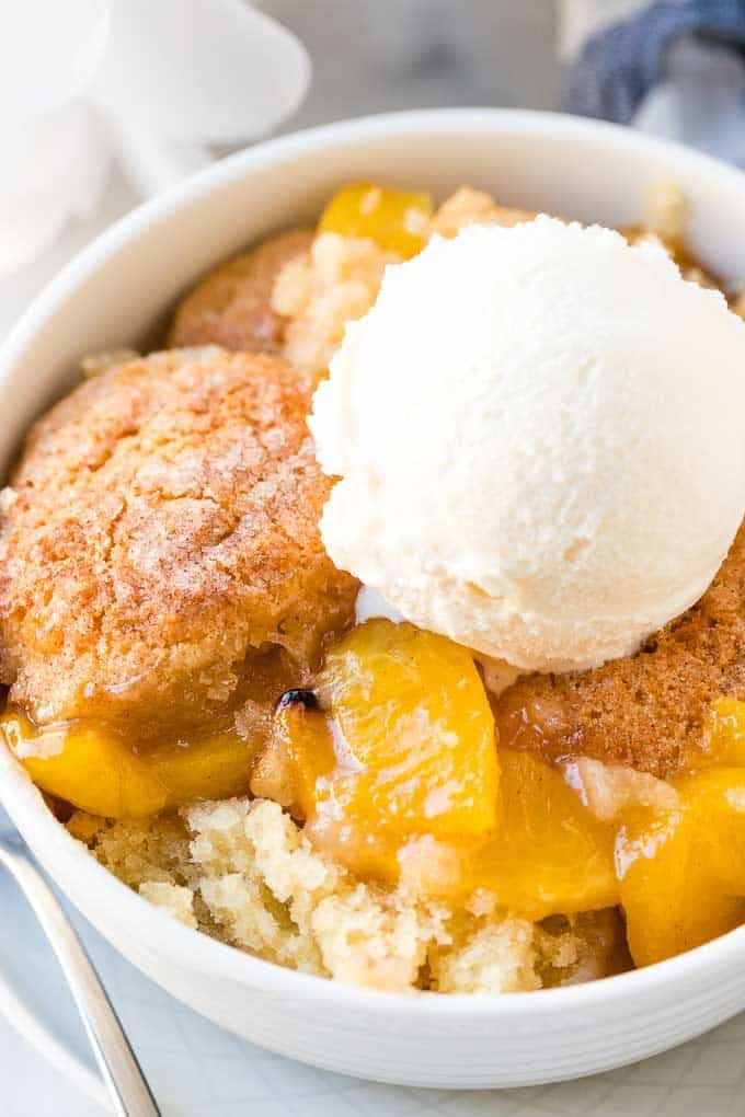 Close-up of a bowl of peach cobbler in a white bowl with a scoop of vanilla ice cream on top.