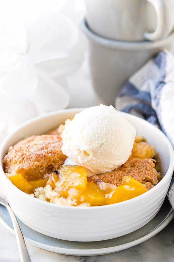 A bowl of peach cobbler in a white bowl with a scoop of vanilla ice cream on top.