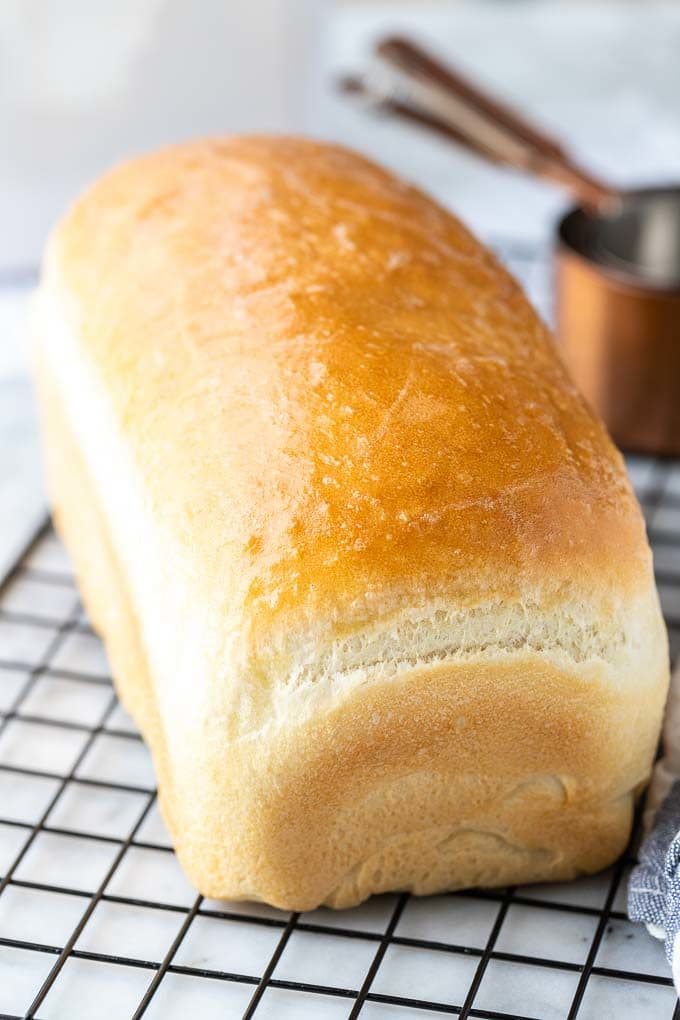 A loaf of white bread on a black cooling rack next to some bronze measuring cups