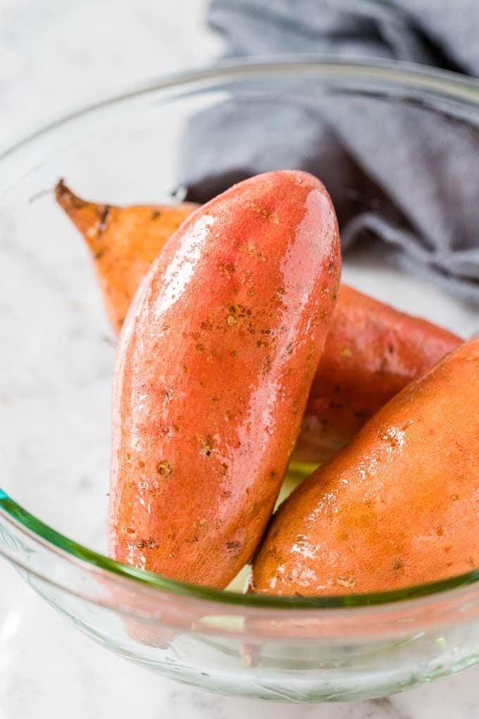 A glass bowl with raw, whole sweet potatoes, covered with olive oil