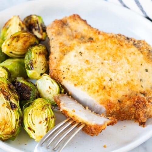 A breaded pork chop on a plate next to Brussels sprouts.