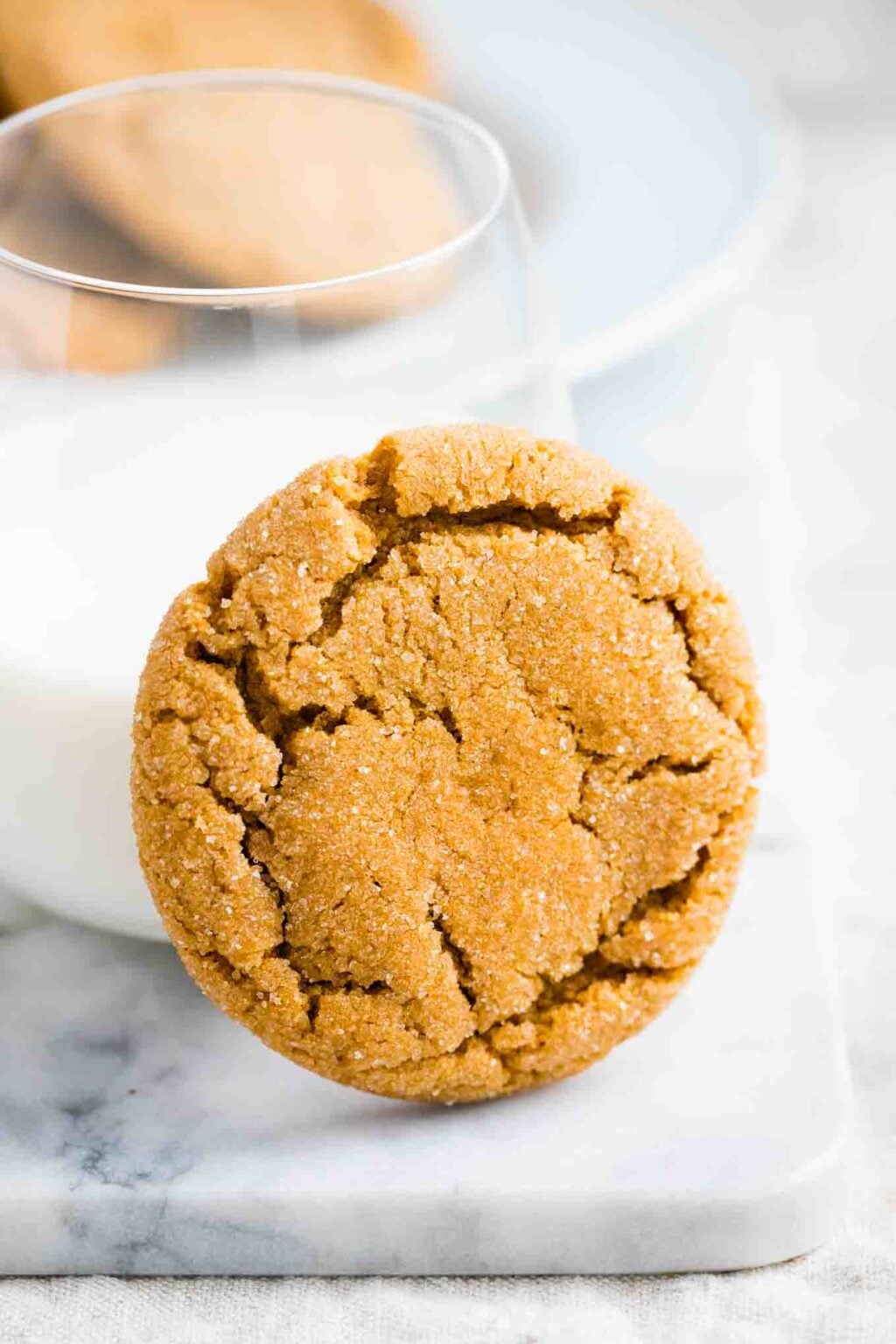 A sugar coated ginger snap cookie with a crackled top leaning against a glass of milk on a white marble board