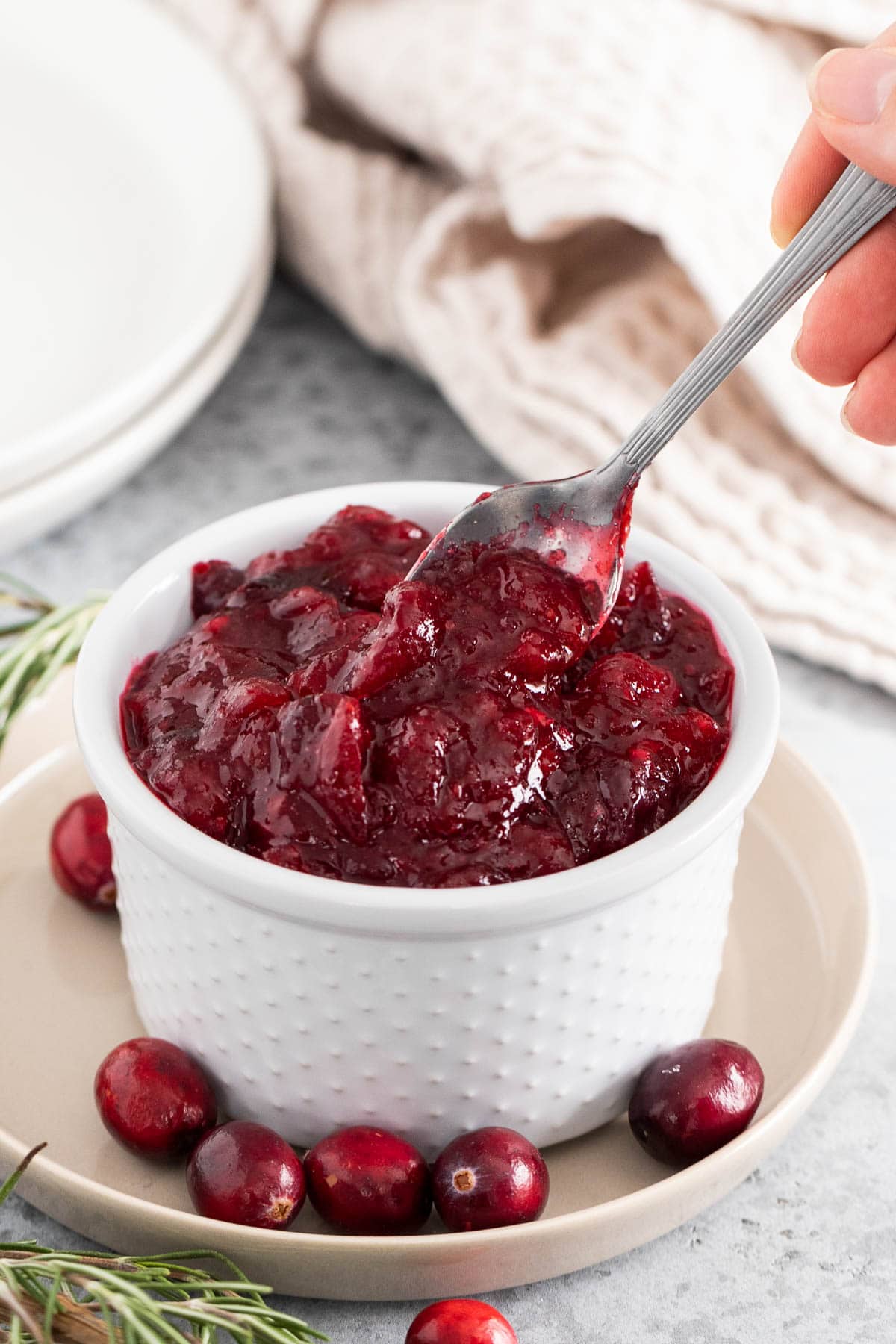 A woman's hand holding a spoon scooping out some cranberry sauce of a white bowl.