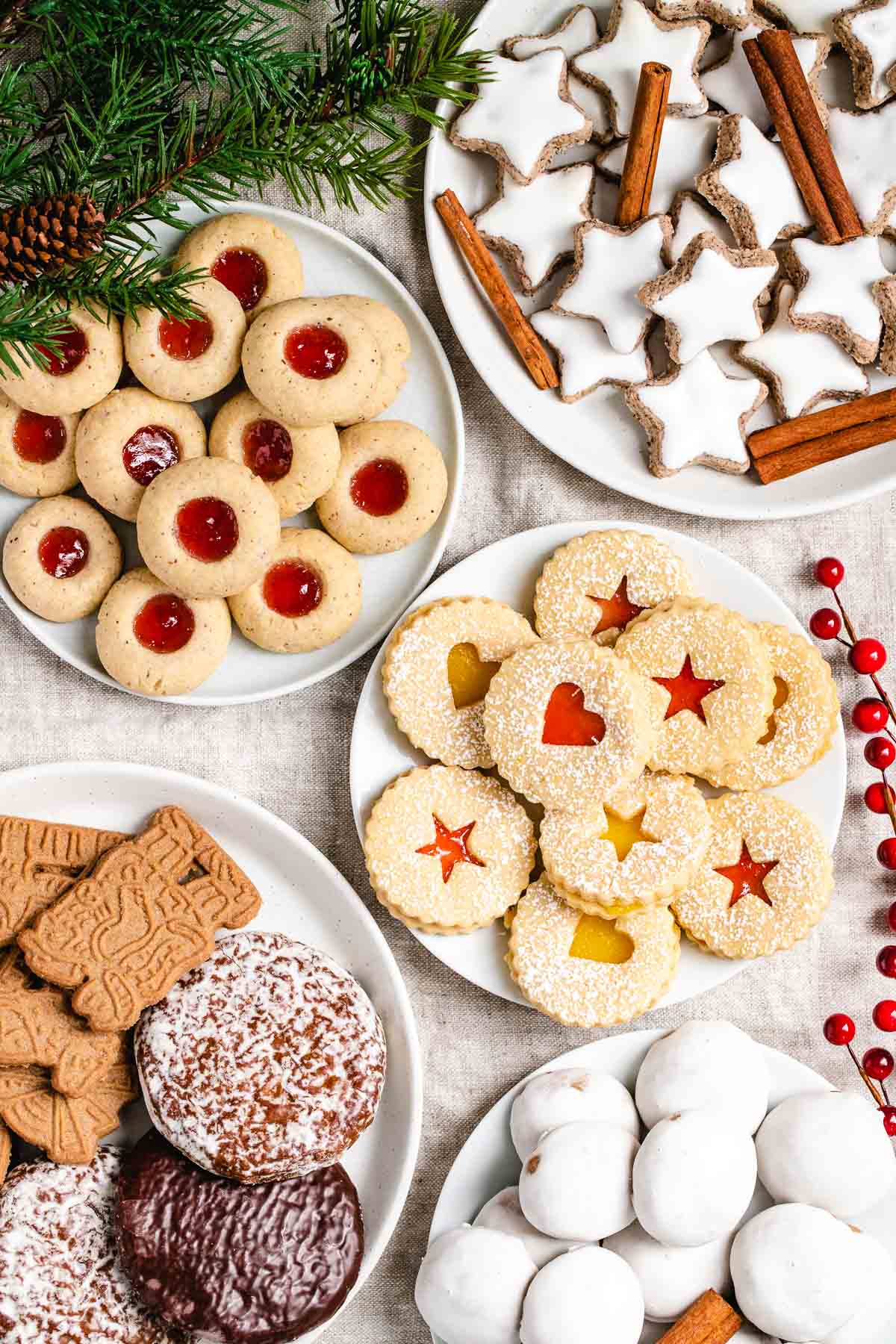 A collection of German Christmas cookies on various plates garnished with pine branches and red berries. There are Cinnamon stars, garnished with cinnamon sticks, thumbprint cookies with strawberry jam, linzer cookies with strawberry jam and lemon curd, Gingerbread and spekulatius cookies and Pfeffernuesse.