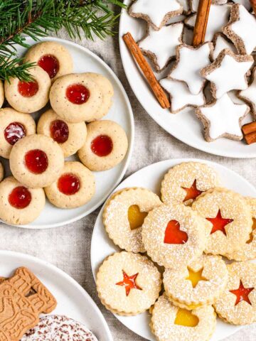 A collection of German Christmas cookies on various plates garnished with pine branches and red berries. There are Cinnamon stars, garnished with cinnamon sticks, thumbprint cookies with strawberry jam, linzer cookies with strawberry jam and lemon curd and spekulatius cookies.
