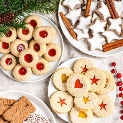 A collection of German Christmas cookies on various plates garnished with pine branches and red berries. There are Cinnamon stars, garnished with cinnamon sticks, thumbprint cookies with strawberry jam, linzer cookies with strawberry jam and lemon curd and spekulatius cookies.