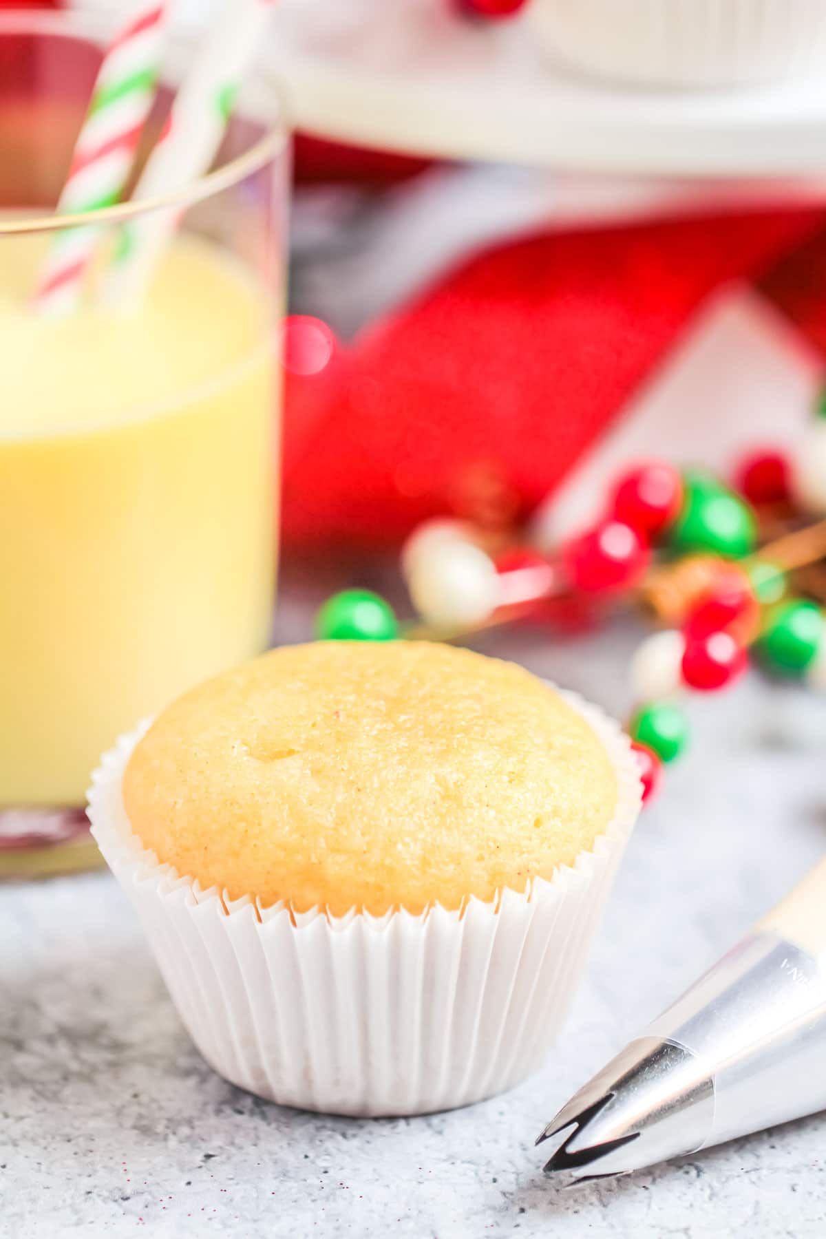 An unfrosted cupcake next to a piping bag, in the background a glass of eggnog with a straw in it.