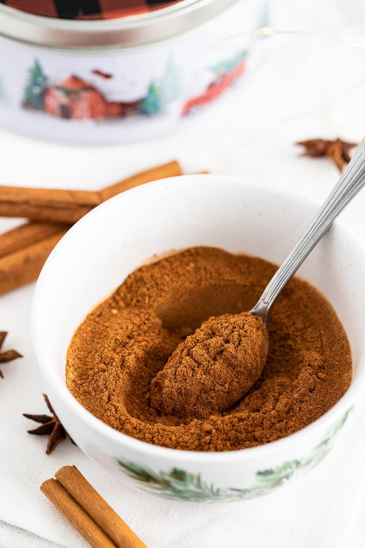 A white bowl filled with ground spices and a spoon in it, next to cinnamon sticks and star anise.