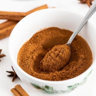 A white bowl filled with ground spices and a spoon in it, next to cinnamon sticks and star anise