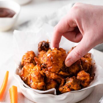 A female hand holding a cauliflower wing coated in BBQ sauce next to a white bowl filled with more wings