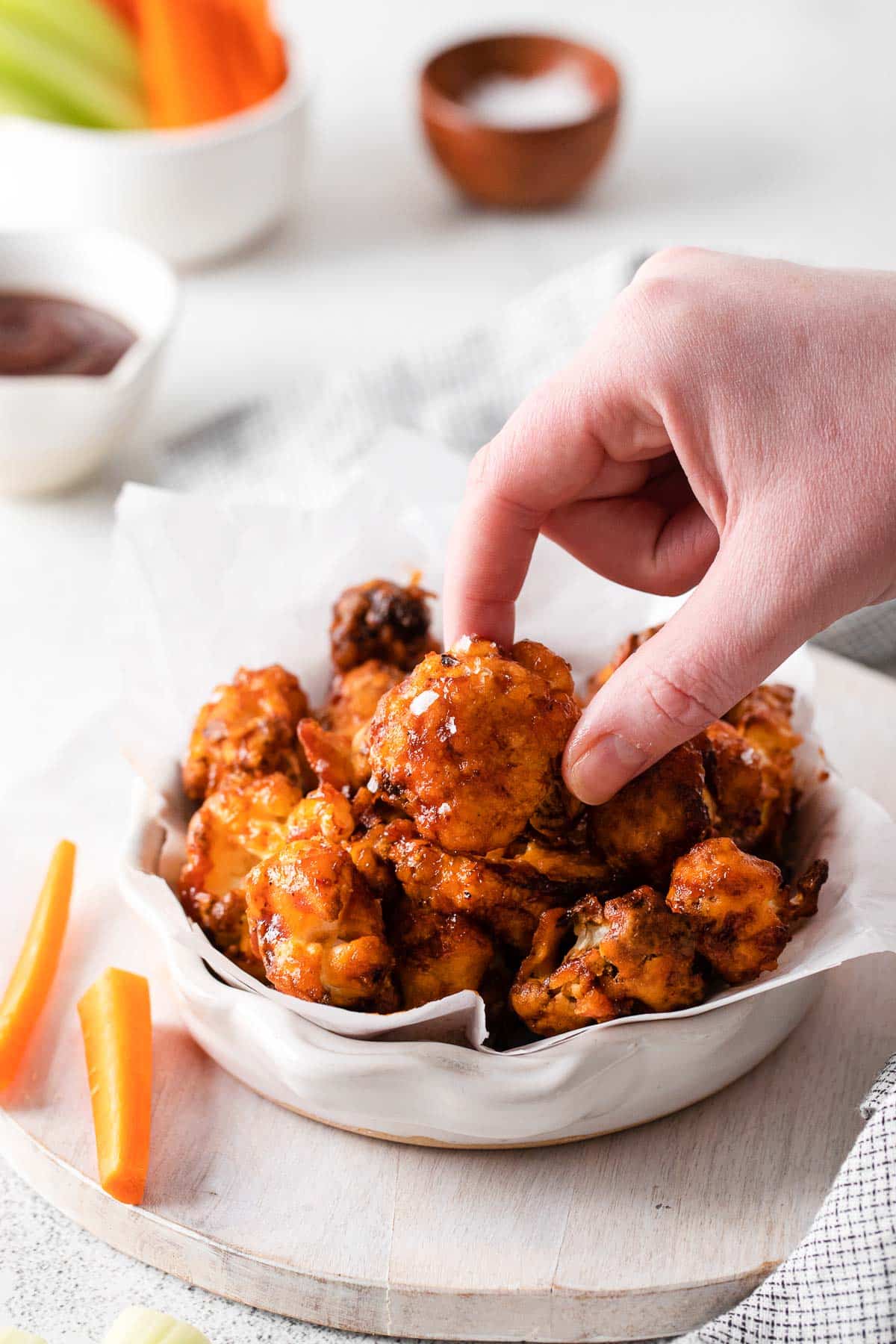 A female hand holding a cauliflower wing coated in BBQ sauce next to a white bowl filled with more wings