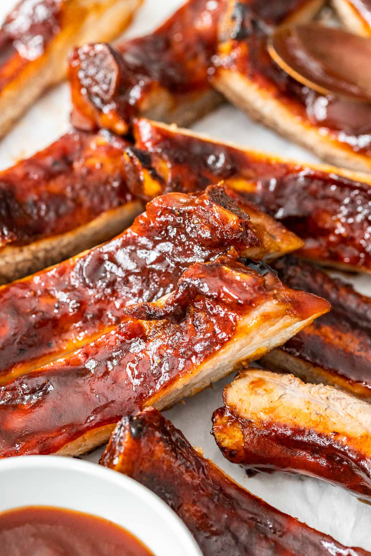 sliced ribs slathered with BBQ sauce on parchment paper next to a small white bowl with BBQ sauce