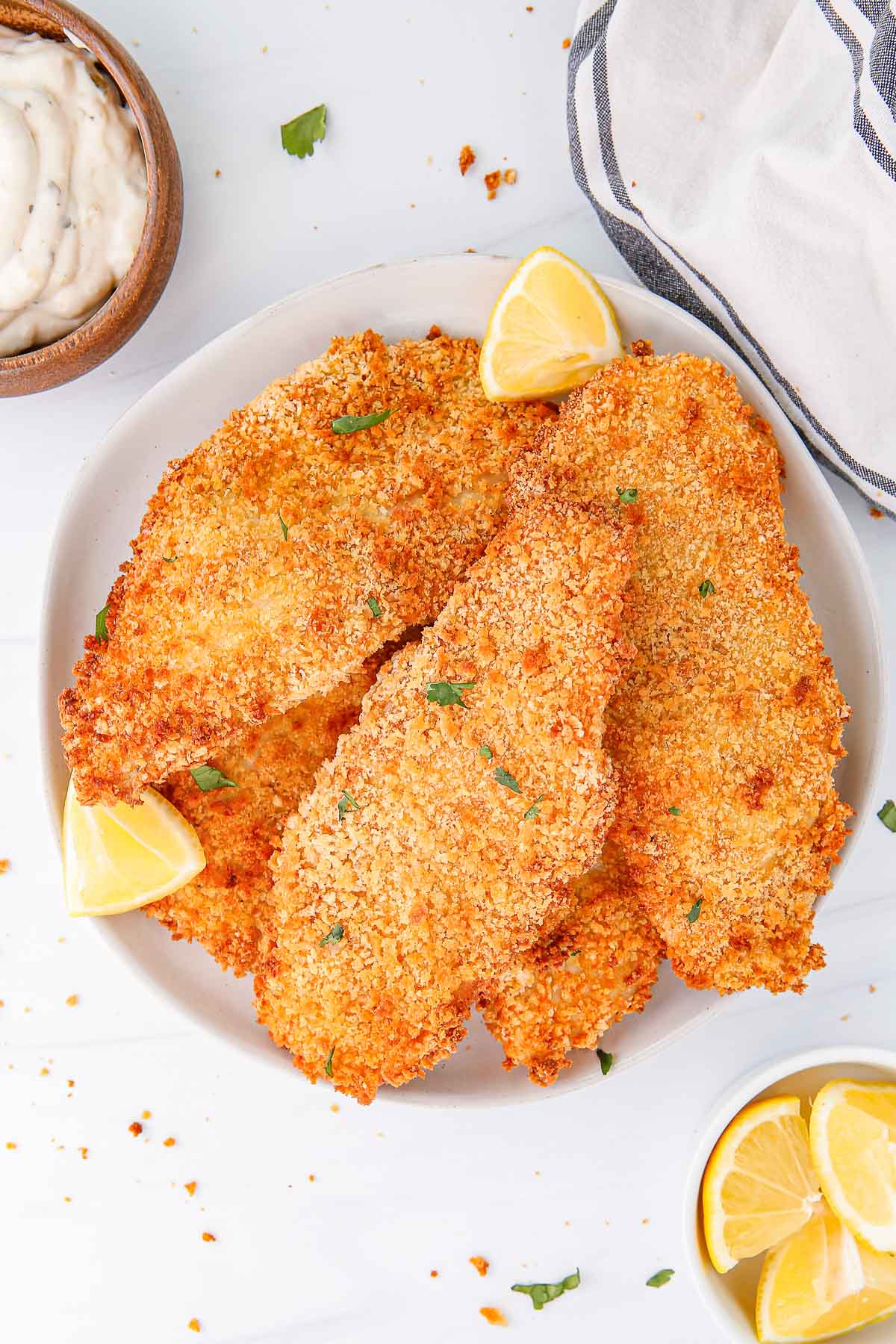 a white plate with air fried breaded fish fillets with lemon wedges, next to a bowl of lemon wedges and a bowl of mayonnaise