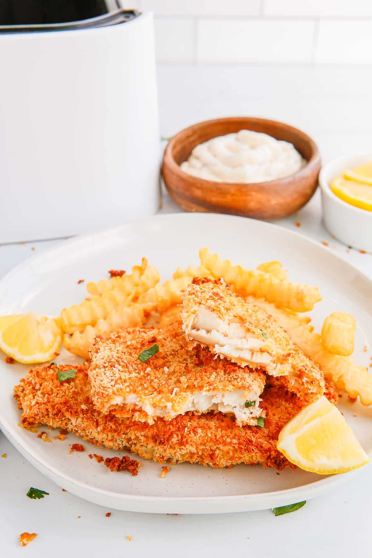 breaded fish fillets with fries and lemon wedges in front of a bowl of mayonnaise, a bowl of lemon wedges and an air fryer basket