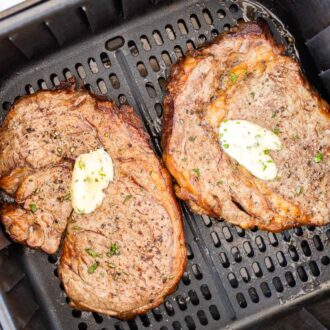 Two cooked steaks with a dollop of garlic butter on top in a black air fryer basket.