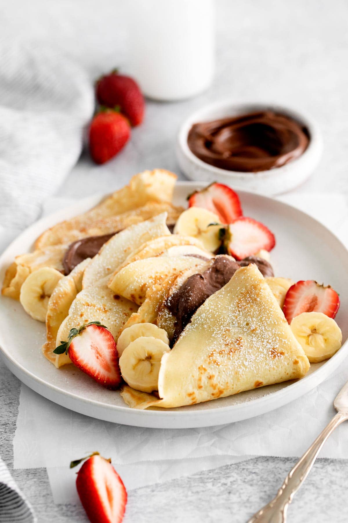 French crepes on a white plate garnished with strawberries, banana slices, and Nutella