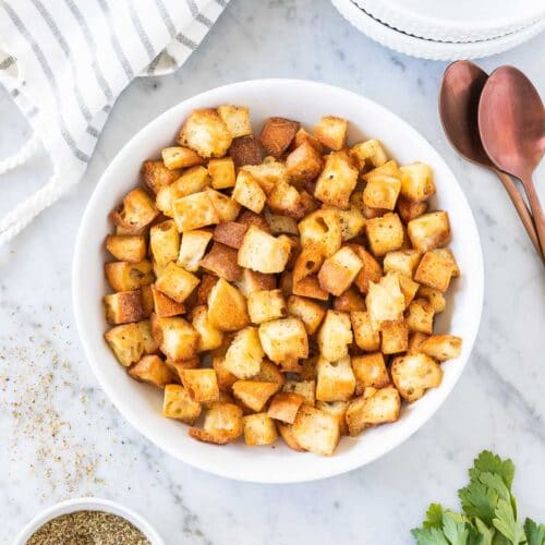 Croutons in a white bowl next to a spoon and a kitchen towel on a marble board.