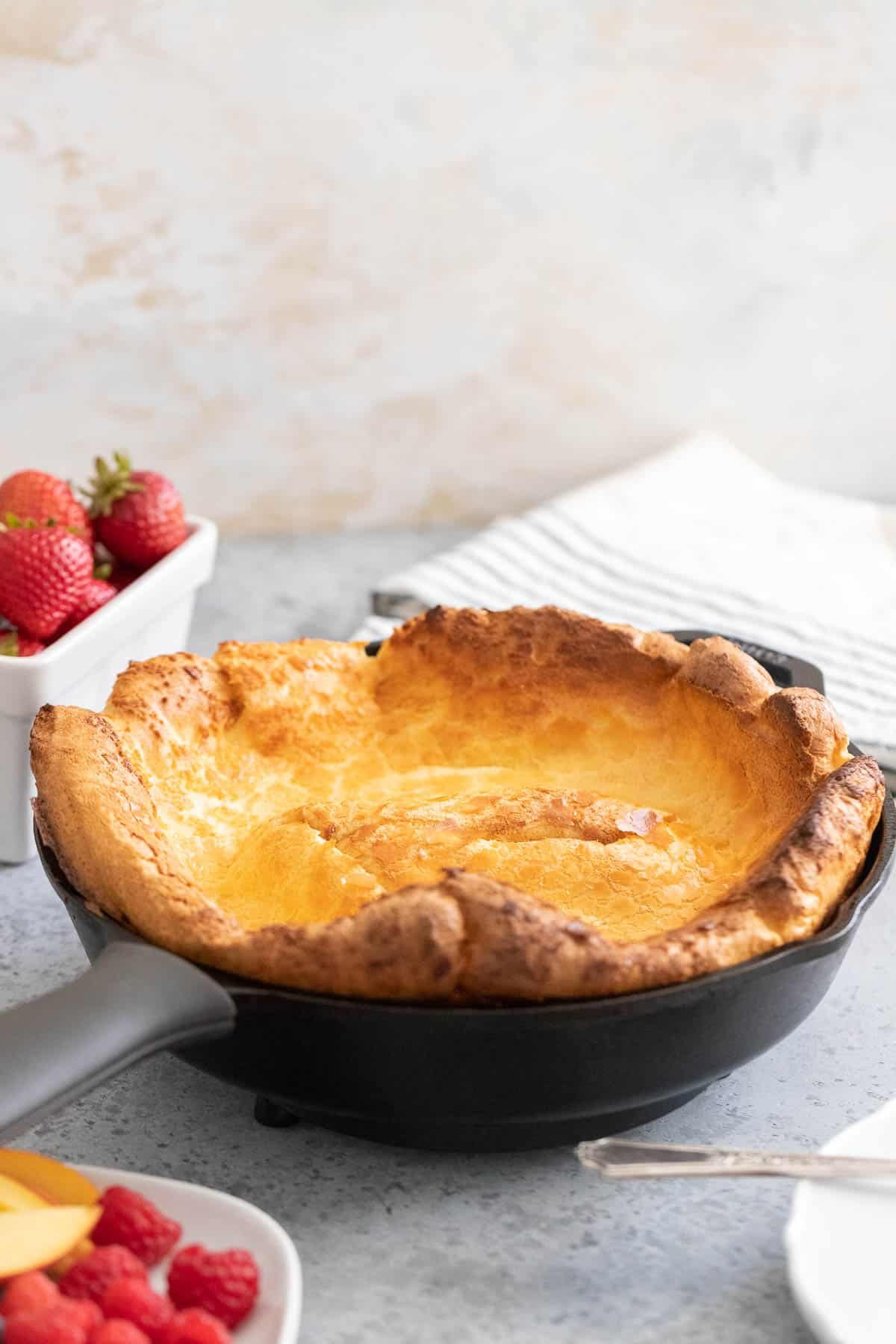 A Dutch Baby pancake in a cast iron pan next to a bowl of strawberries.