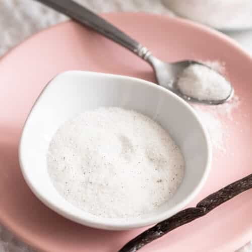 A small bowl filled with vanilla sugar on a pink plate with a spoon and vanilla bean next to it.