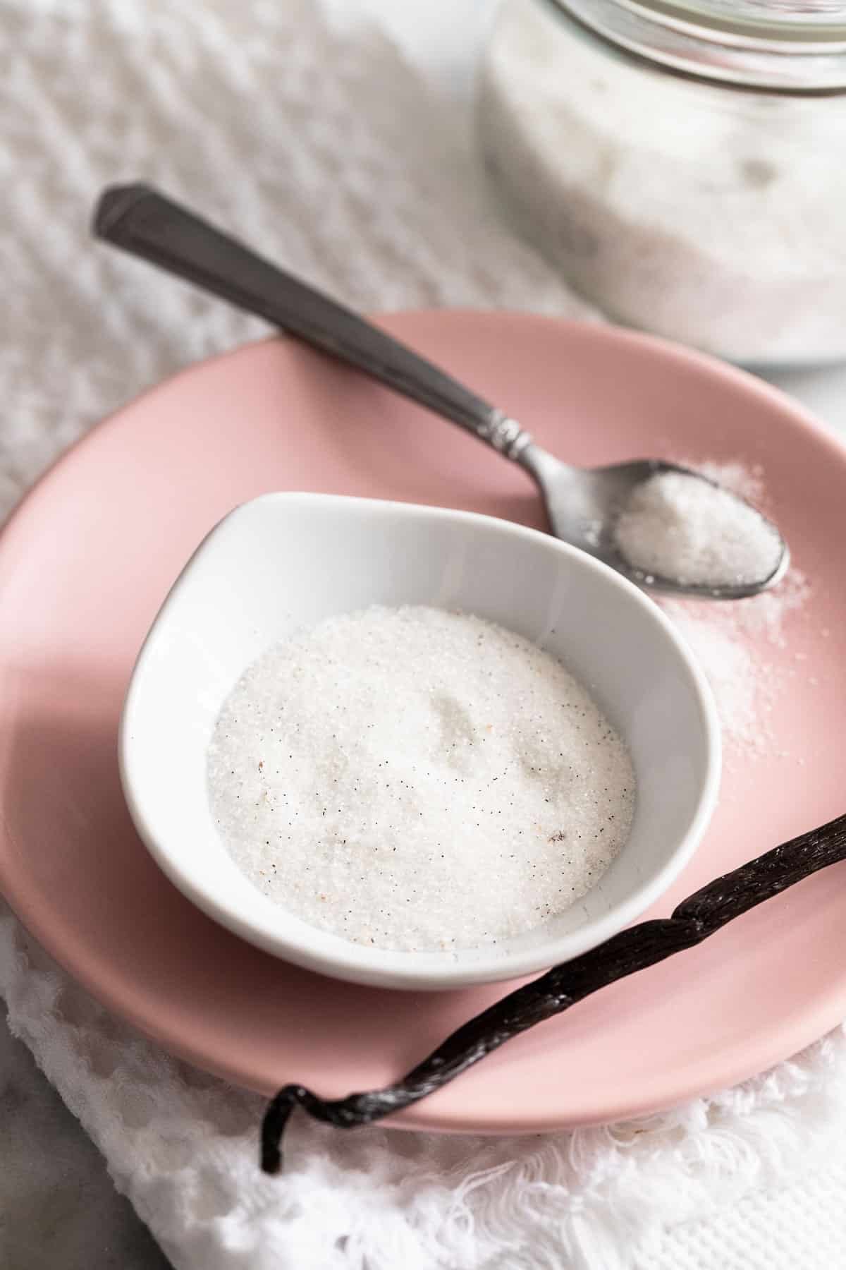 A small bowl filled with vanilla sugar on a pink plate with a spoon and vanilla bean next to it.