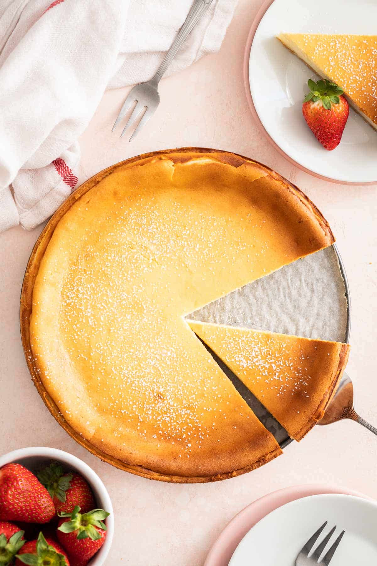 A German Cheesecake on a light pink surface next to strawberries and plates.