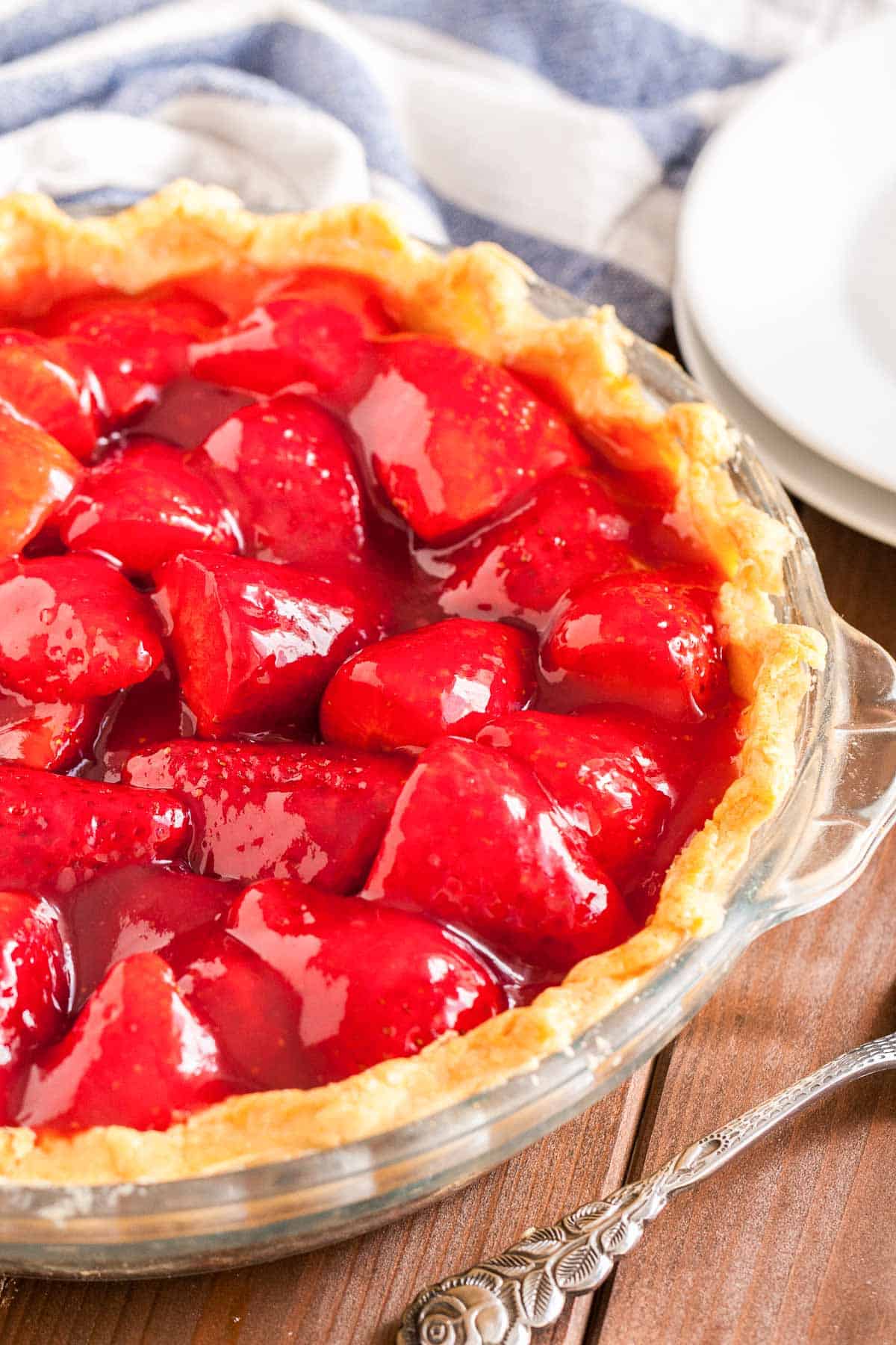 Close-up of a glass baking dish, with strawberry pie on a brown wood table, a white and blue dish towel, 2 stacked white plates and a cake server.