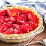 Close-up of a glass baking dish, with strawberry pie on a brown wood table, a white and blue dish towel, 2 stacked white plates and a cake server.