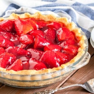 Close-up of a glass baking dish, with strawberry pie on a brown wood table, a white and blue dish towel, 2 stacked white plates and a cake server.